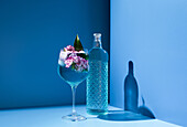 Transparente Glasflasche mit alkoholischem Getränk auf Tisch neben frischem Cocktail mit Eis und Blumen vor blauem Hintergrund