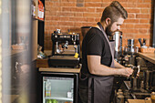 Seitenansicht eines bärtigen männlichen Barista, der einen Krug mit einer Serviette säubert, während er am Tresen neben der Kaffeemaschine steht und in einem Café arbeitet