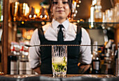 Long spoon placed on glass cup with ice and lime on blurred background of young female bartender in pub