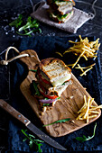 Draufsicht auf ein leckeres Sandwich mit Tomate, Zwiebel, Käse und Fleisch auf einem Holzbrett mit Pommes frites und Messer auf einem blauen Tisch