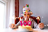 Positive aged female in sunglasses and with glowing Bengal lights sitting at table with cake with 100 candle and celebrating anniversary while looking at camera