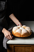 High angle of crop unrecognizable baker standing at counter with fresh bread buns placed on baking pan in kitchen