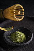 Spoon with dried matcha tea leaves on black tableware with chasen for traditional oriental ceremony