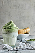 Glass of fresh dalgona coffee with matcha and Japanese tea ceremony accessories consisting of bamboo whisk called chasenand straw placed on wooden table