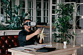 Unrecognizable male chef in uniform sitting at table with various documents and smartphone and holding grilled corn while experiencing virtual reality in VR headset in restaurant