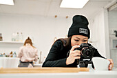 Ethnic female in beanie taking photo of bowl on digital camera against unrecognizable partner in kitchen