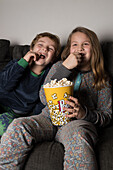 Young cheerful siblings kids sitting comfortable on sofa with popcorn bucket while eating and watching TV