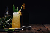 Glass of alcoholic cocktail decorated with pineapple piece and leaves with paper straw on tray near shaker and bottle with shot glass at table on black background