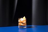 Glass of alcoholic cocktail made with red vermouth served with orange slices in light studio with blue and black wall