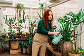 Caring red haired female with jug watering plants growing in pots placed on shelf in indoor garden with various houseplants