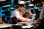 Young cooks men in aprons carefully serving meal in white ceramic dishes on restaurant kitchen