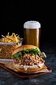 Appetitlicher Burger mit Pulled Pork, Barbecue-Soße, Cheddar-Käse und Salatmischung, serviert mit Pommes frites und einem Glas kaltem Bier auf einem Holzbrett