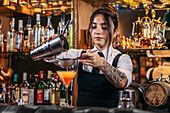 Young tattooed brunette pouring alcohol from shaker through sieve while making tequila sunrise cocktail during work in bar