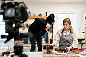 Unrecognizable woman taking photo of chocolate muffins on digital camera against blogger talking during cooking process in kitchen