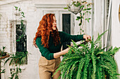 Serious female with ginger hair trimming green branch of potted fern growing in indoor garden