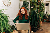Konzentrierte junge Freiberuflerin sitzt auf einem Stuhl und surft auf ihrem Netbook zu Hause mit Topfpflanzen bei der Fernarbeit