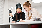 Cheerful female blogger showing cellphone to ethnic girlfriend with professional photo camera while interacting at home