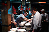 Young cooks men in aprons carefully serving meal in white ceramic dishes on restaurant kitchen