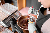 From above soft focus of anonymous worker using infrared thermometer to measure temperature of liquid chocolate in bowl in confectionery