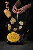 Crop-Koch schlägt Ei auf, während eine halbe geschälte Kartoffel mit rohen Stücken auf das Omelett in der Bratpfanne tropft