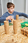 Child sitting at table with pile of cardboard cups and containers against gardening shovel and fork while looking away