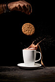 Crop anonymous person throwing tasty sweet cookies into white ceramic cup with splashing coffee on table in kitchen on black background