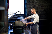 Junger fröhlicher Mann in grauer Schürze rührt in einer großen Metallpfanne in einer Restaurantküche