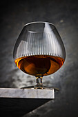 Fragile transparent snifter glass with alcoholic brown cognac placed on corner of marble table against gray background in light studio