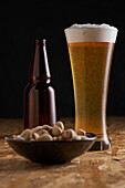 Glass of fresh cold beer placed on wooden table near bottle and plate with pistachios