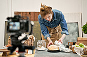 Young female blogger with fresh wheat bread against photo camera during cooking process at table in house