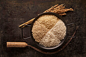 From above of bowl with assorted types of dried rice placed near bunch of ears and sickle on black background