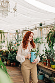 Charming female with long ginger hair standing with jug of seedling in indoor garden with various potted houseplants at home