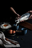 Crop view of hands of anonymous woman under dramatic lighting preparing ingredients like milk, flour and eggs in order to make a dough