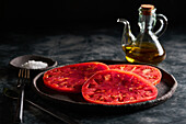 Delicious sliced tomatoes in cast iron plate near sea salt and jug of olive oil on concrete table