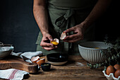 Unrecognizable guy in apron breaking raw egg over bowl while preparing pastry on lumber table near kitchenware