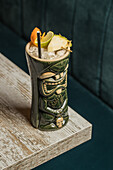 From above of large sculptural tiki cup filled with booze decorated with straw and fruits placed on green rug against wooden table
