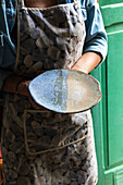 Crop unrecognizable artisan in apron demonstrating creative ceramic plate in workshop