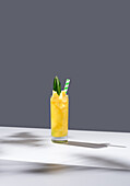 Glass full of cold pineapple juice with green leaves and straw placed on sunlit table against gray background