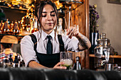 Young brunette in uniform mixing cold cocktail in glass cup while standing behind counter and working in bar