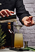 Crop unrecognizable barman pouring cocktail through strainer from shaker in glass filled with ice cubes against brick wall