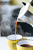 Freshly brewed steaming hot coffee pouring from metal moka pot into yellow cup on table in light kitchen on blurred background