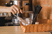 Crop anonymous male barista taking black plastic straw from cup using metal tongs in cafe