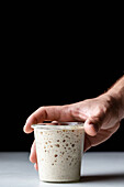 Hand of crop anonymous person demonstrating white round glass cup with sourdough starter and covered with glass lid placed on table against black background