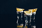 Tequila shots with salt and lemon placed on reflective surface against dark background