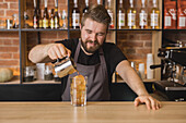 Cheerful bearded male barista in apron pouring ingredient from stainless pitcher into glass with ice cubes in coffee house