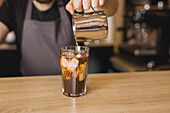 Crop male barista in apron adding milk into glass with iced coffee in coffee house during working day