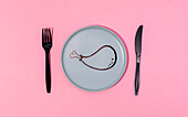 Top view of black plastic fork and knife placed on pink table on gray plate with painted cartoon chicken drumstick