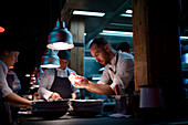 Young cooks men in aprons carefully serving meal in white ceramic dishes on restaurant kitchen