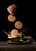 Crop anonymous person throwing tasty sweet cookies into green ceramic cup with splashing coffee on table in kitchen on black background