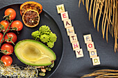 Top view of decorative inscription near plate with fresh half of avocado and bundle of cherry tomatoes near red orange slices